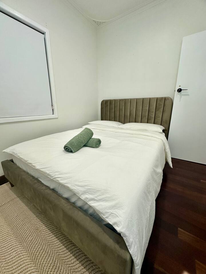 Camera da letto pulita e semplice con un letto a due piazze, struttura imbottita marrone chiaro, lenzuola bianche e asciugamani verdi. Presenta pareti bianche, una porta bianca, pavimenti in legno scuro e una luce naturale soffusa per un'atmosfera calda e accogliente.