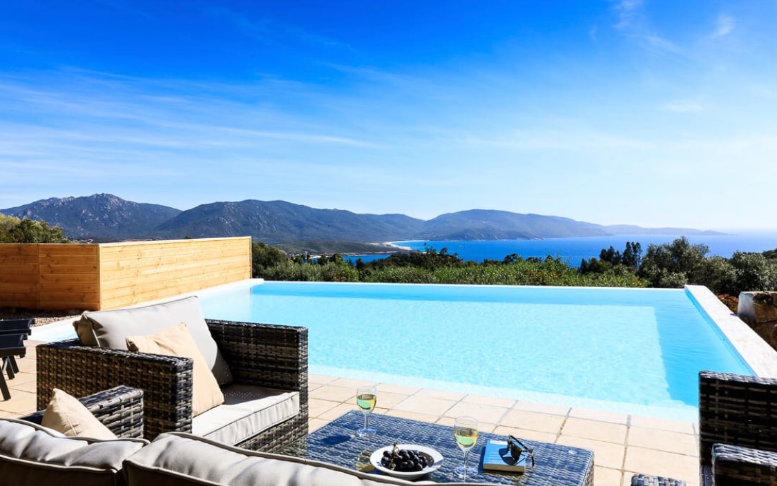 An inviting outdoor seating area features a comfortable arrangement of wicker furniture facing a large infinity pool. Expansive views of rolling hills and the ocean stretch in the background under a clear blue sky, enhancing the relaxing ambiance.