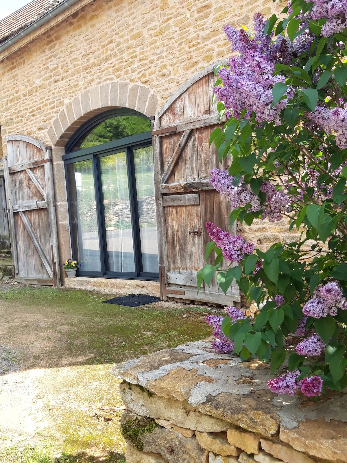 An entrance to the renovated barn is framed by rustic wooden doors, complementing the natural stone facade. Lilac flowers bloom nearby, adding a touch of color to the inviting exterior. Sunlight filters through the glass doors, enhancing the tranquil outdoor setting.