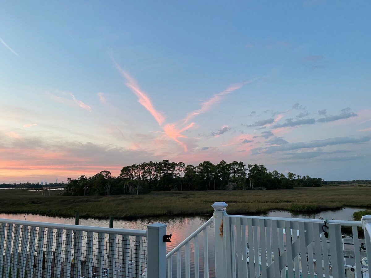 A serene view of the estuary is captured at sunset, with soft pink hues in the sky contrasting against green tree lines in the background. The calm water reflects the colorful clouds, creating a tranquil atmosphere.