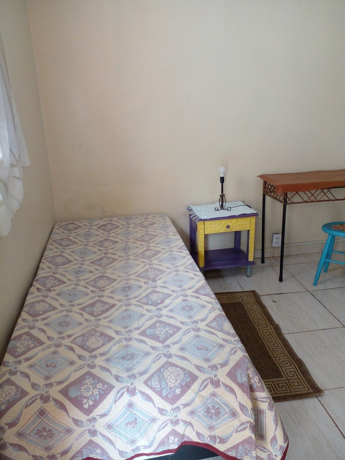 A single bed is positioned against a light-colored wall, covered with a patterned bedspread. A small side table with a lamp sits beside the bed. A wooden desk is visible in the corner of the room, accompanied by a blue chair and a decorative rug on the tiled floor.