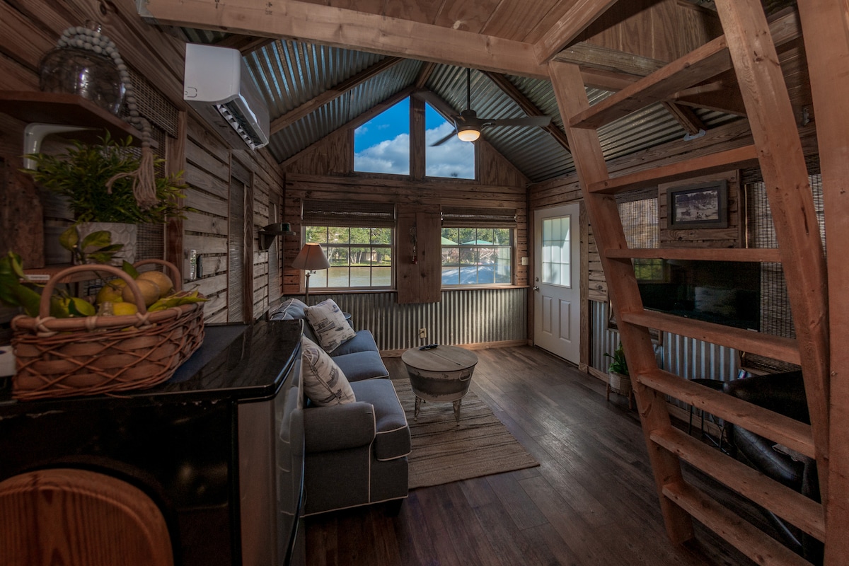 The interior space features rustic wood paneling and metal accents, with a cozy couch positioned beside a small coffee table. A staircase leads to the lofted sleeping area. Large windows allow natural light to fill the room, creating a relaxed ambiance.