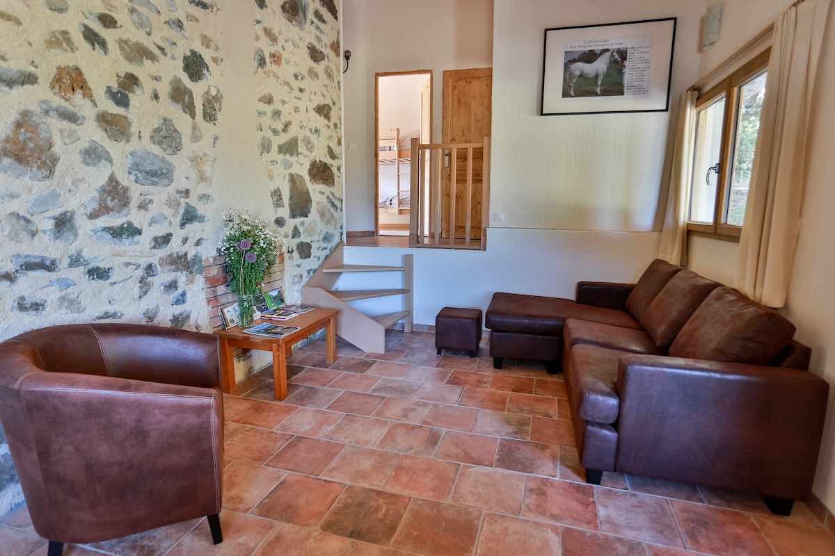 A cozy living area features a stone wall that adds character. Comfortable brown furniture, including a sofa and armchair, is arranged for relaxation. Natural light enters through large windows, illuminating a small dining table. A staircase leads to additional living spaces.