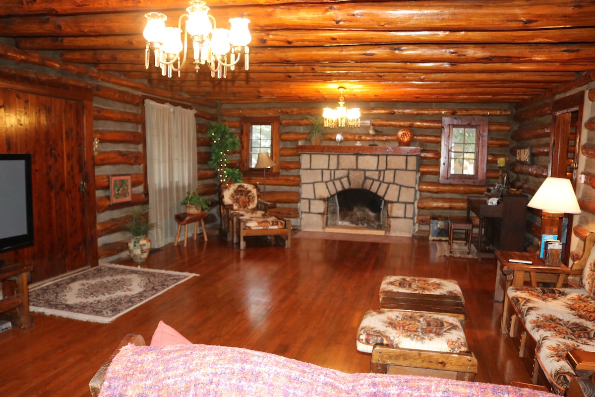 The living area showcases exposed log walls and a warm wooden floor. A stone fireplace serves as a central feature, surrounded by comfortable seating, including floral-patterned chairs. A chandelier provides soft lighting, while a small table and lamp add functional decor adjacent to the fireplace.