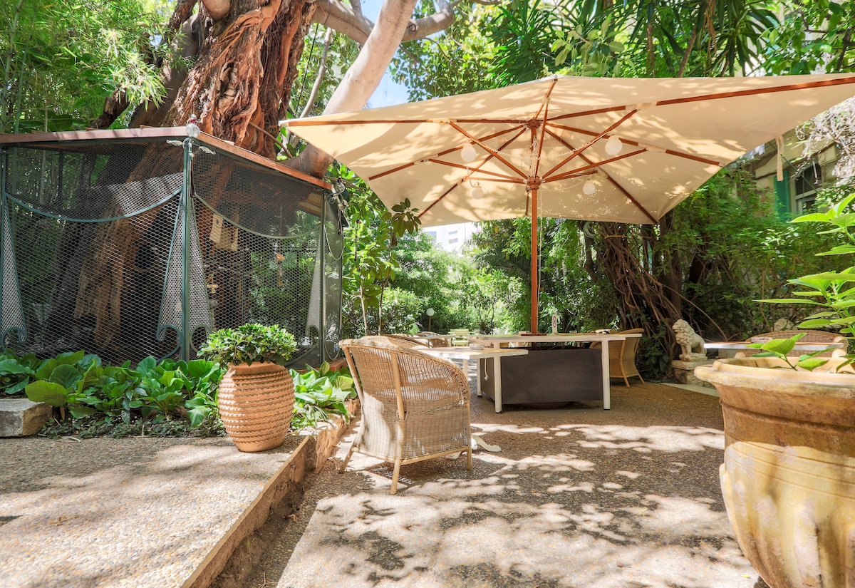 A shaded outdoor seating area is surrounded by lush greenery, featuring a large umbrella over a wooden table and chairs. A tropical tree stands nearby, with a subtle path leading through the garden, creating a serene retreat.