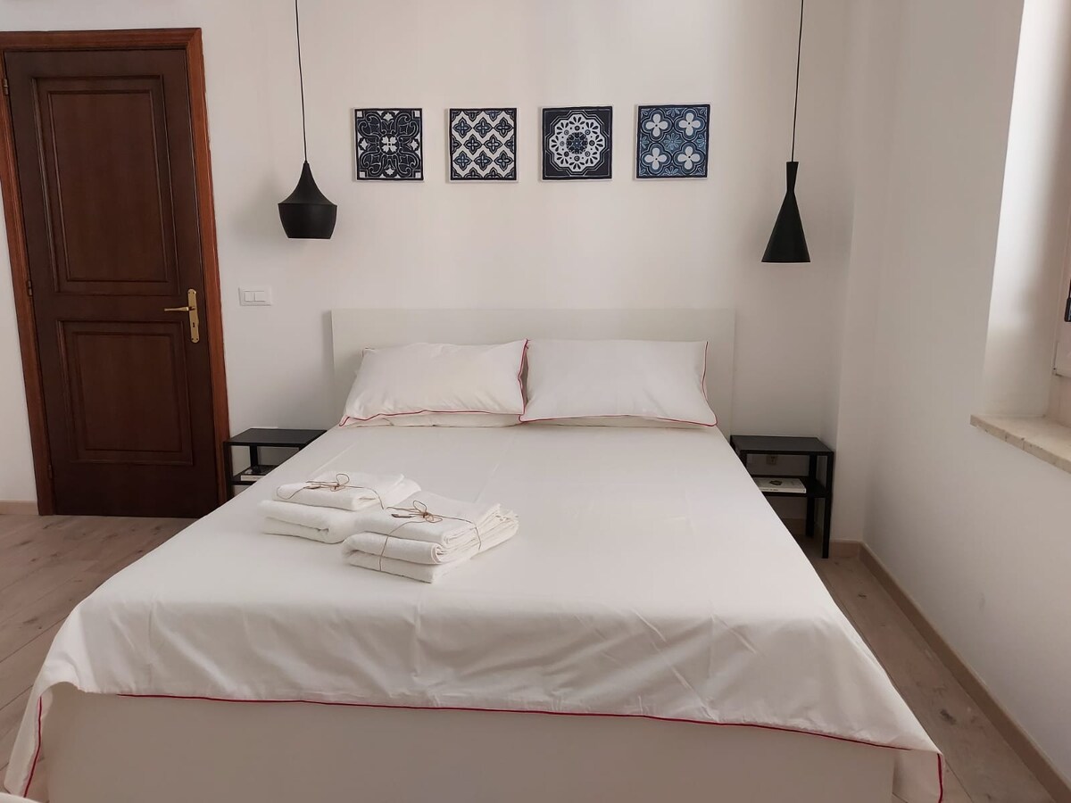 A spacious bedroom features a large bed with crisp white linens and neatly arranged towels. Two minimalist black pendant lights hang above, while decorative wall art in coordinating colors adorns the wall. A wooden door provides access to the room, complemented by a window allowing natural light.
