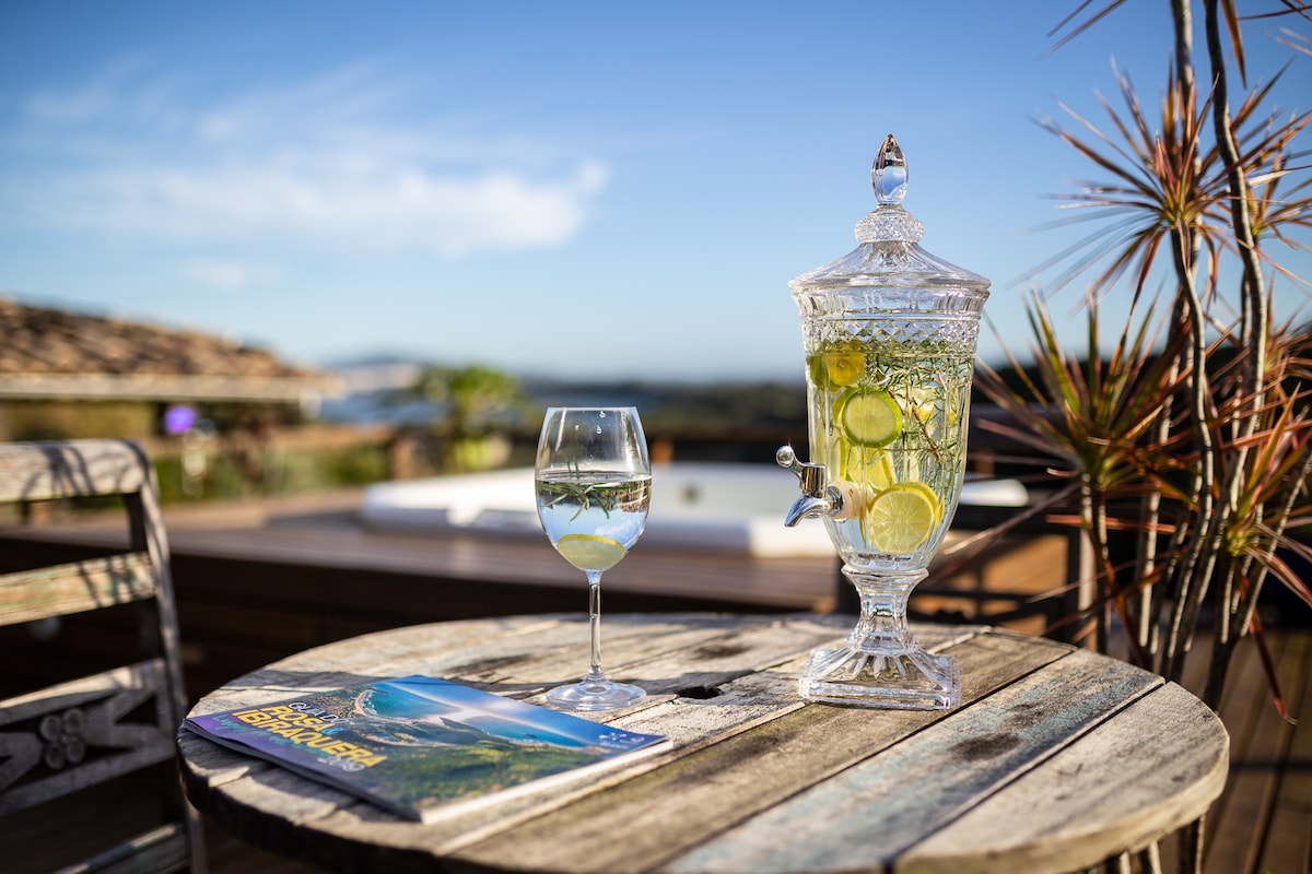 A refreshing beverage is displayed in a crystal dispenser with slices of lemon. An elegantly styled glass sits on a rustic wooden table, accompanied by a travel magazine. Soft natural light illuminates the scene, revealing distant scenic views.