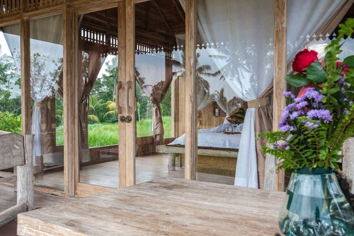 A natural wooden structure features large glass doors that open to a spacious interior. Soft, flowing drapes frame the entrance, with a view of a gently made bed visible inside. A simple wooden table is adorned with a vase of fresh flowers.
