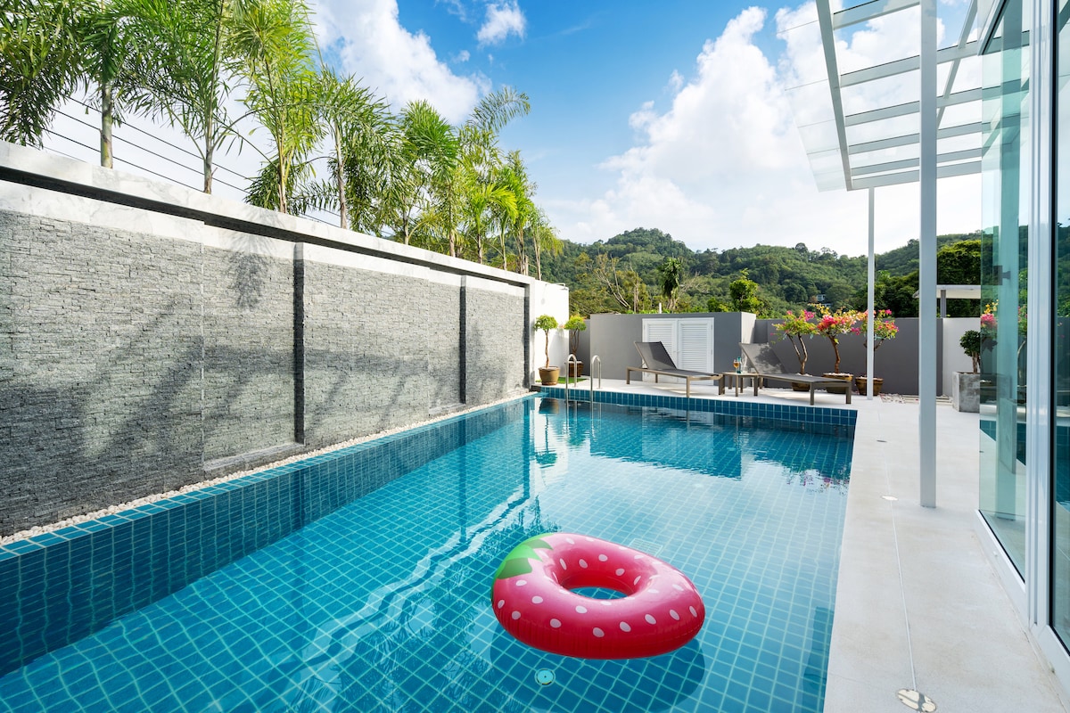 A private pool is surrounded by lush greenery and stone walls, reflecting clear blue skies. A colorful inflatable float is positioned in the center of the water. Lounge chairs are placed nearby, providing a relaxing space to enjoy the serene environment.