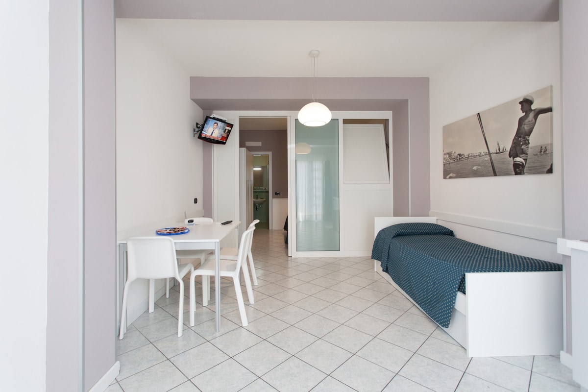 A bright living space features a cozy bed adorned with a blue coverlet. A white dining table with four chairs occupies one corner, while a wall-mounted television is visible. Light-toned walls and tiled flooring enhance the open atmosphere, with a thoughtful arrangement of furnishings.