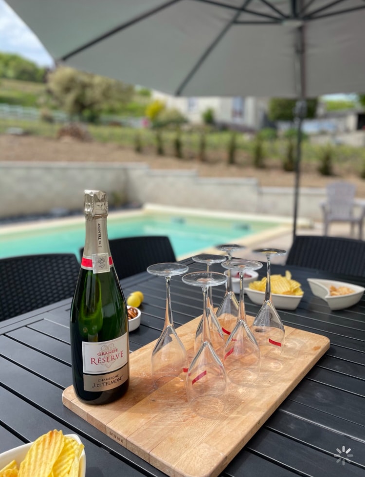 A serving board holds a bottle of champagne and six elegant glasses in a relaxed outdoor dining setting. In the background, a private pool is visible, along with snacks of chips and olives, all under a large umbrella.