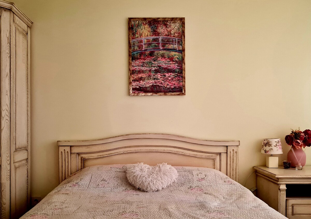 A serene bedroom showcases a spacious bed adorned with a decorative quilt. A heart-shaped pillow rests atop the bed. The wall displays a colorful artwork of a bridge over a floral scene, while a nightstand features a lamp and a vase of flowers.