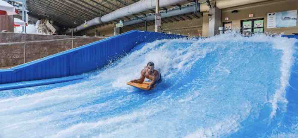 A person is riding a flowboard on a water slide, surrounded by cascading blue water. The scene captures the excitement of indoor water activity, with the slide featuring a steep incline and a smooth surface. The background includes the entry area and some structures typical of a water park.