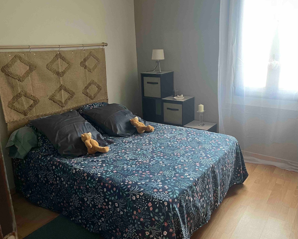 A cozy bedroom features a spacious bed with a floral-patterned cover and two plush toys resting on the pillows. Soft lighting is provided by a bedside lamp, and a unit with dark storage boxes adds to the decor. Natural light streams in through a nearby window.