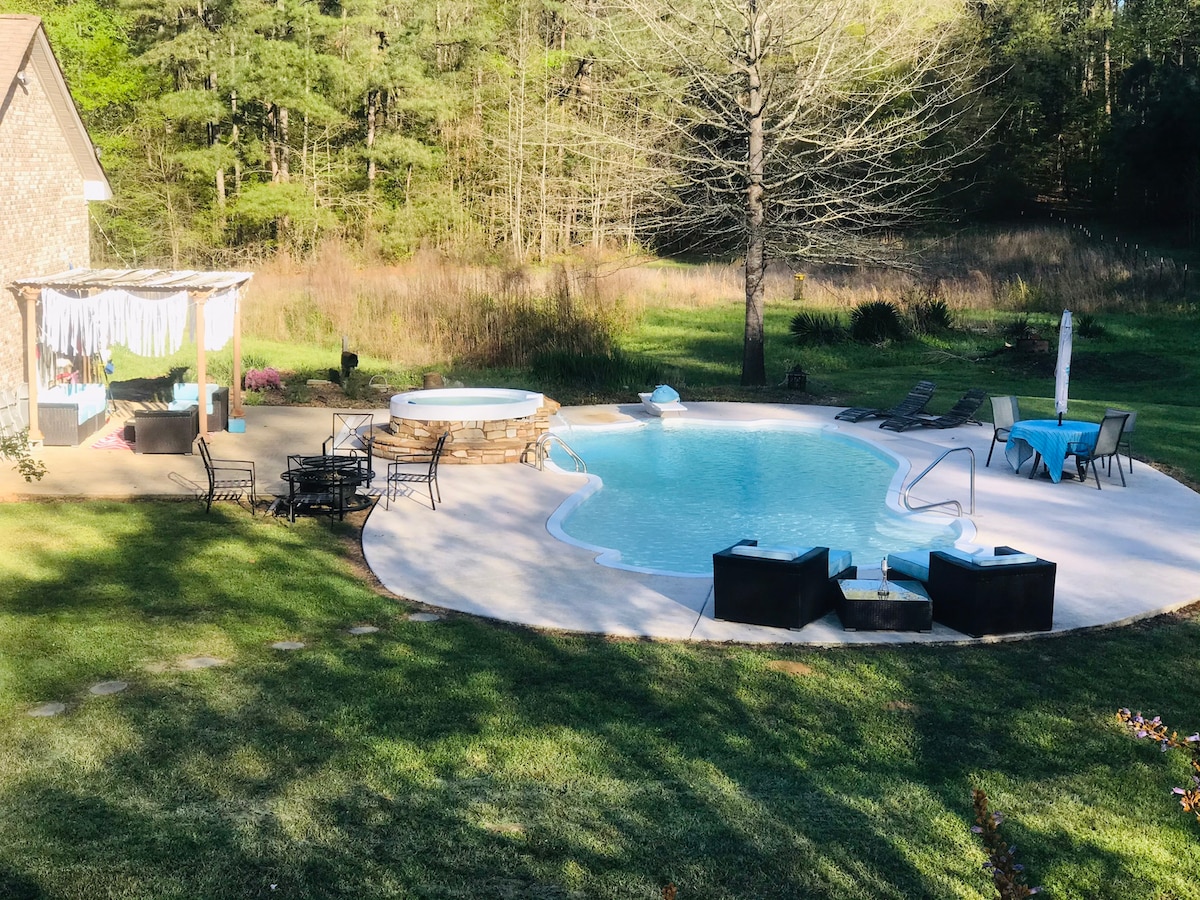 An outdoor pool area features a clear blue swimming pool surrounded by a spacious deck. Lounge chairs and seating are arranged near the pool, while a hot tub provides a relaxing space. Lush greenery and trees encircle the area, creating a serene backdrop.
