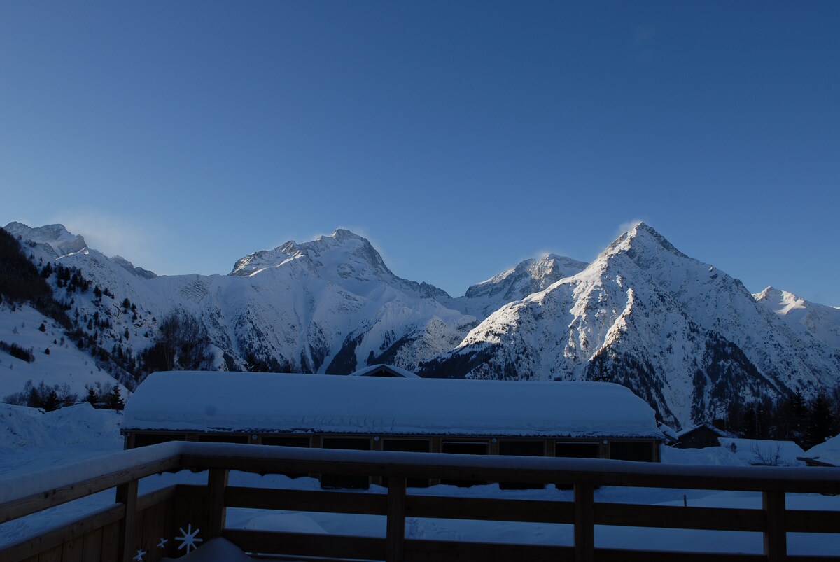 Snow-covered mountains stretch across the horizon under a clear blue sky. The peaceful scene showcases the natural beauty of the alpine landscape, with undulating peaks and valleys offering a serene backdrop. A wooden railing at the foreground provides a perspective of the view from the terrace.