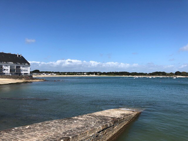 A coastal view showcases calm blue waters extending towards a sandy beach, framed by a distant shoreline. The sky is clear with a few fluffy clouds, and a low stone pier juts into the water, providing a serene focal point.