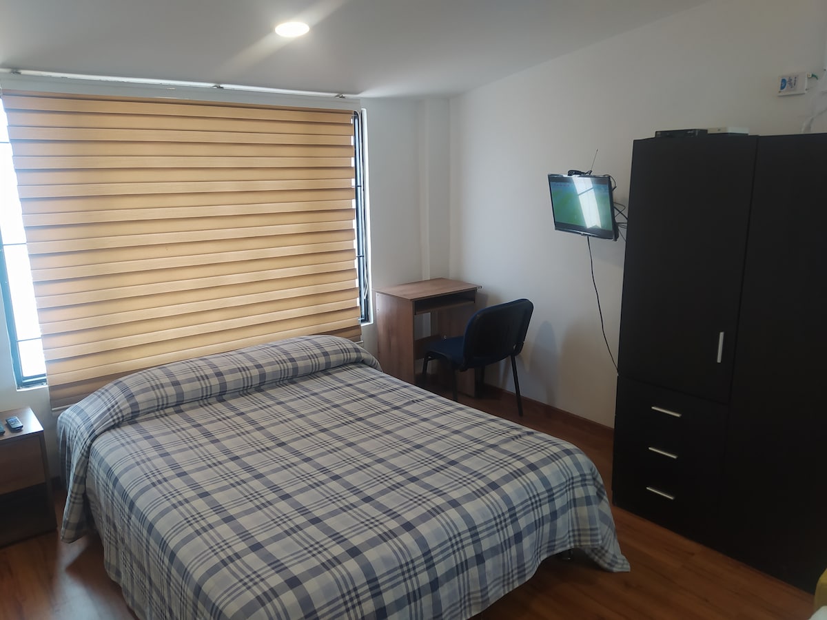 The spacious bedroom features a double bed with a checkered blue and white coverlet, complemented by bedside tables. A desk and chair are situated beside a large window with horizontal blinds. A television is mounted on the wall near a dark wardrobe.
