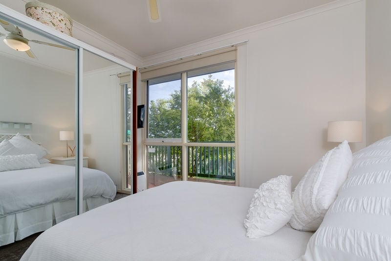 A well-lit bedroom features a spacious bed with white linens and decorative pillows. A wall of mirrored closets reflects the natural light, while a large window offers views of greenery outside. Soft lamps provide an ambient glow, enhancing the airy atmosphere of the room.
