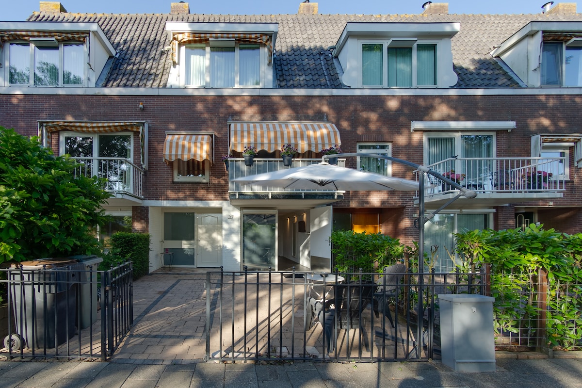 The guesthouse features a welcoming facade with a brick exterior and large windows, adorned with striped awnings. A paved patio area offers seating options under a shade umbrella, surrounded by green foliage, creating a peaceful outdoor space.