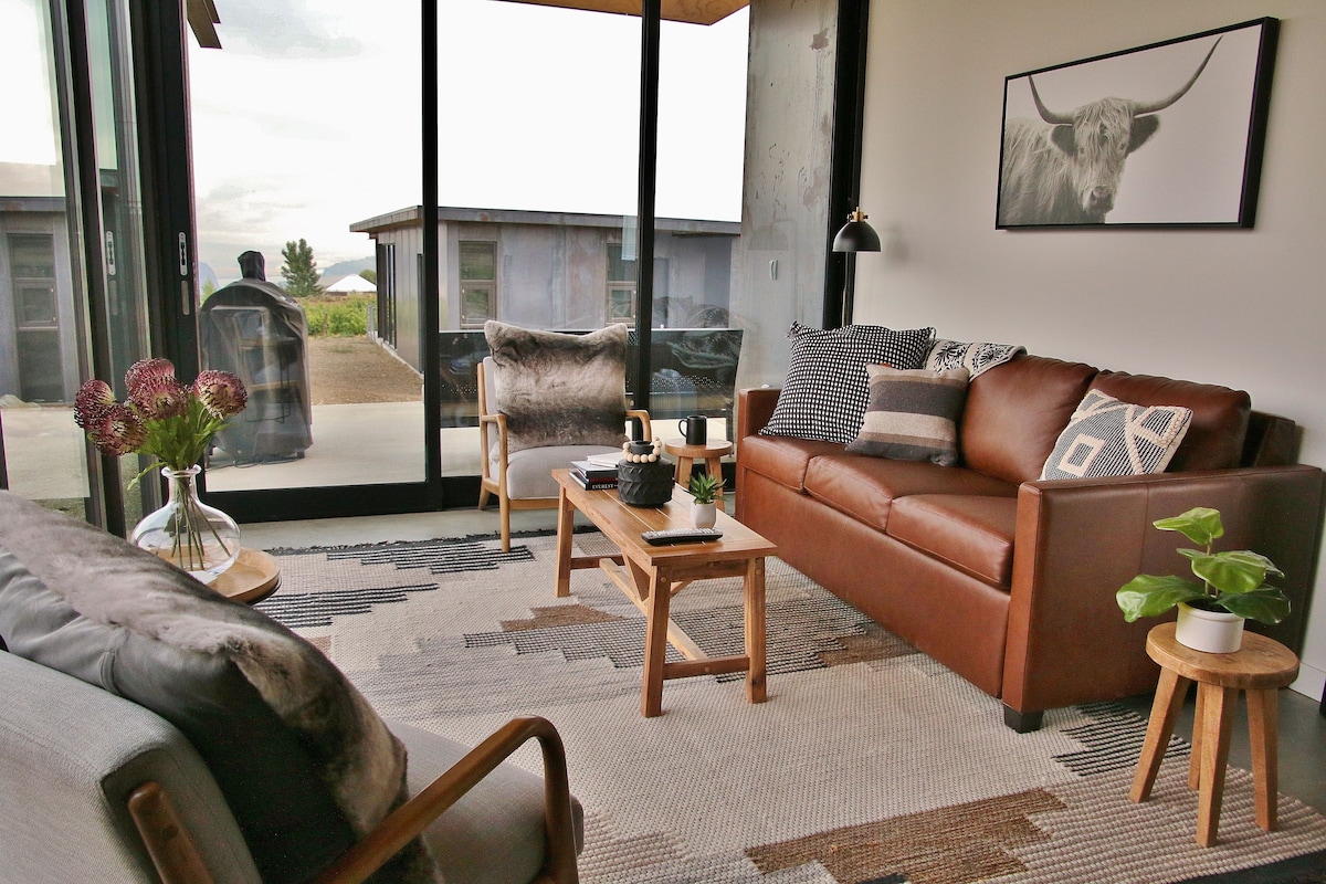A cozy living area is shown with a brown leather sofa and two accent chairs. A woven area rug covers the floor, complementing the wood coffee tables. Large glass doors allow natural light to fill the space, providing a view of the outdoors, framed artwork decorates the walls.