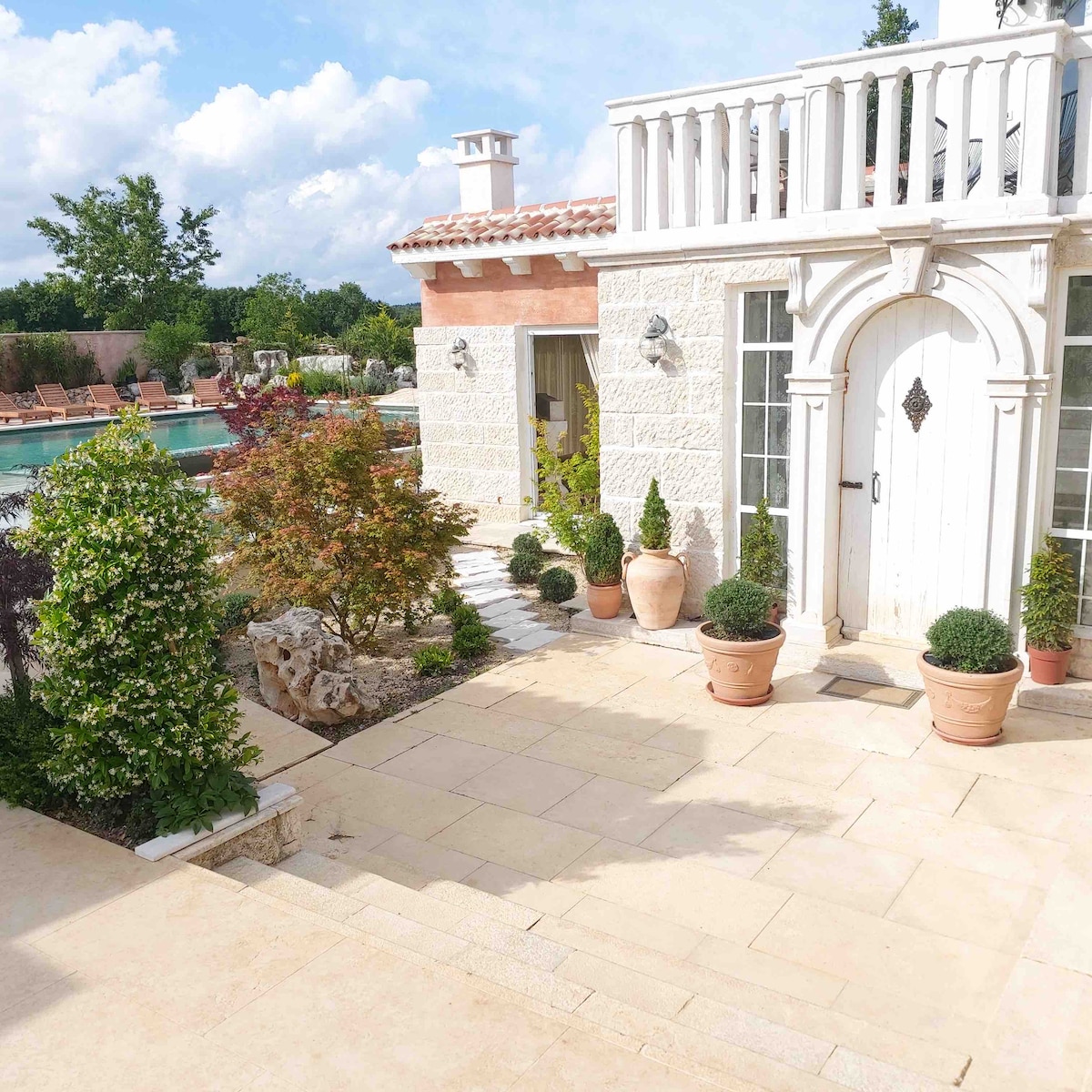 The image displays a stone villa with an arched doorway, flanked by potted plants. A sunlit pathway leads toward a multi-level swimming pool surrounded by neatly arranged landscaping. Lush greenery and a blue sky enhance the serene outdoor setting.