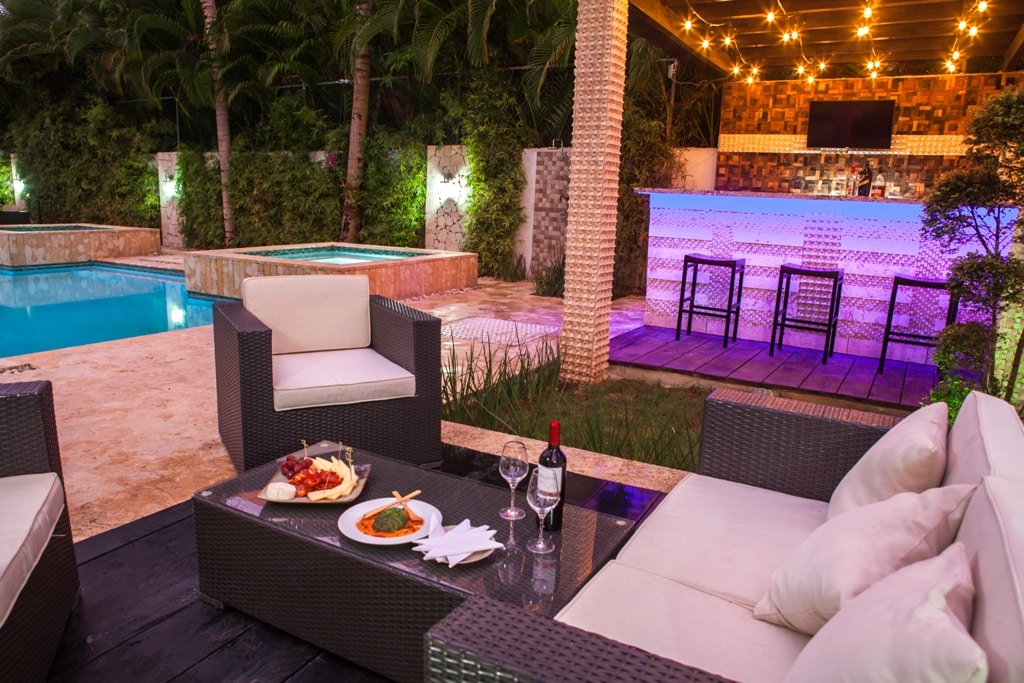 A cozy outdoor seating arrangement features a low table with a plate of food and couple of wine glasses. A pool is visible in the background, along with a bar area illuminated with soft lighting. Lush greenery surrounds the space, creating a secluded atmosphere.