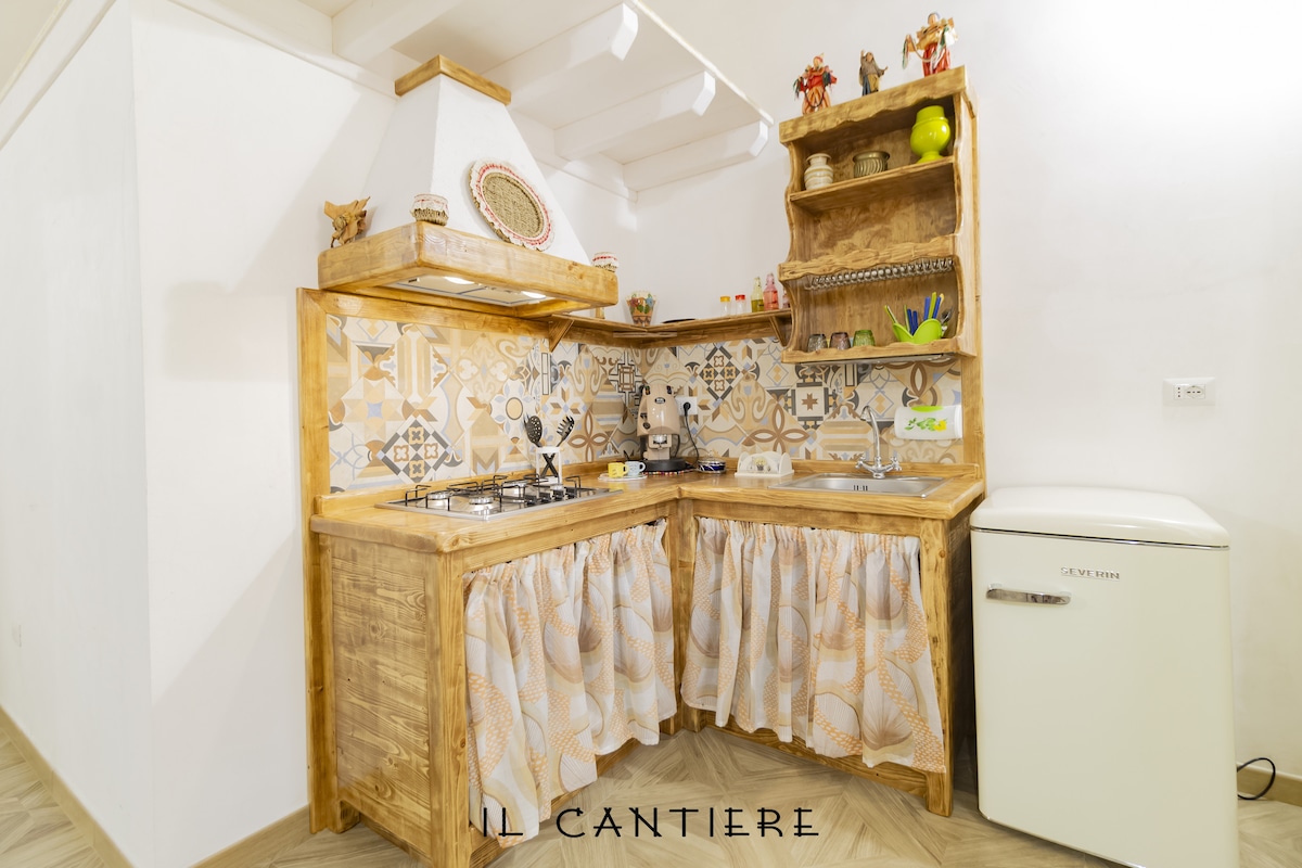 A rustic kitchen corner features a tiled backsplash with decorative patterns. Wooden cabinetry with a cozy curtain drapes beneath the countertop. Appliances, including a vintage-style refrigerator, are seamlessly integrated, ensuring a functional and inviting cooking space.