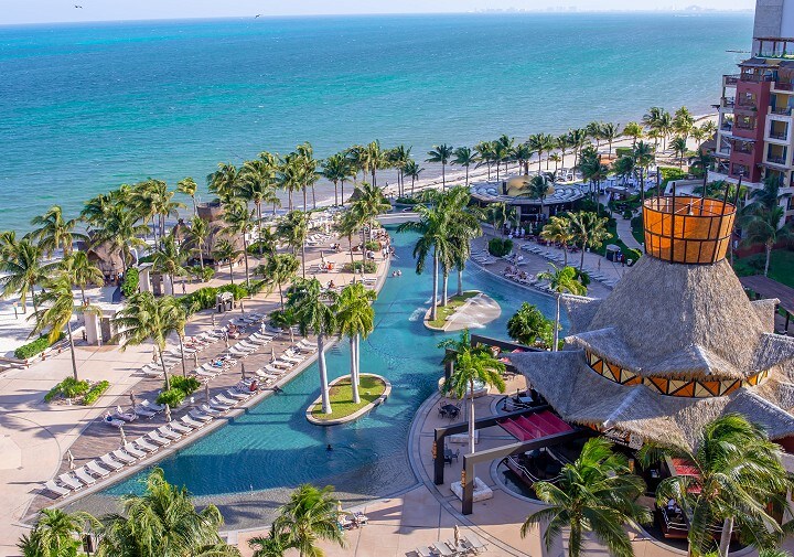 A wide view of the resort reveals manicured tropical landscapes, featuring palm trees surrounding multiple outdoor swimming pools. A thatched-roof structure stands in the foreground, while the azure ocean stretches toward the horizon, offering a serene atmosphere.
