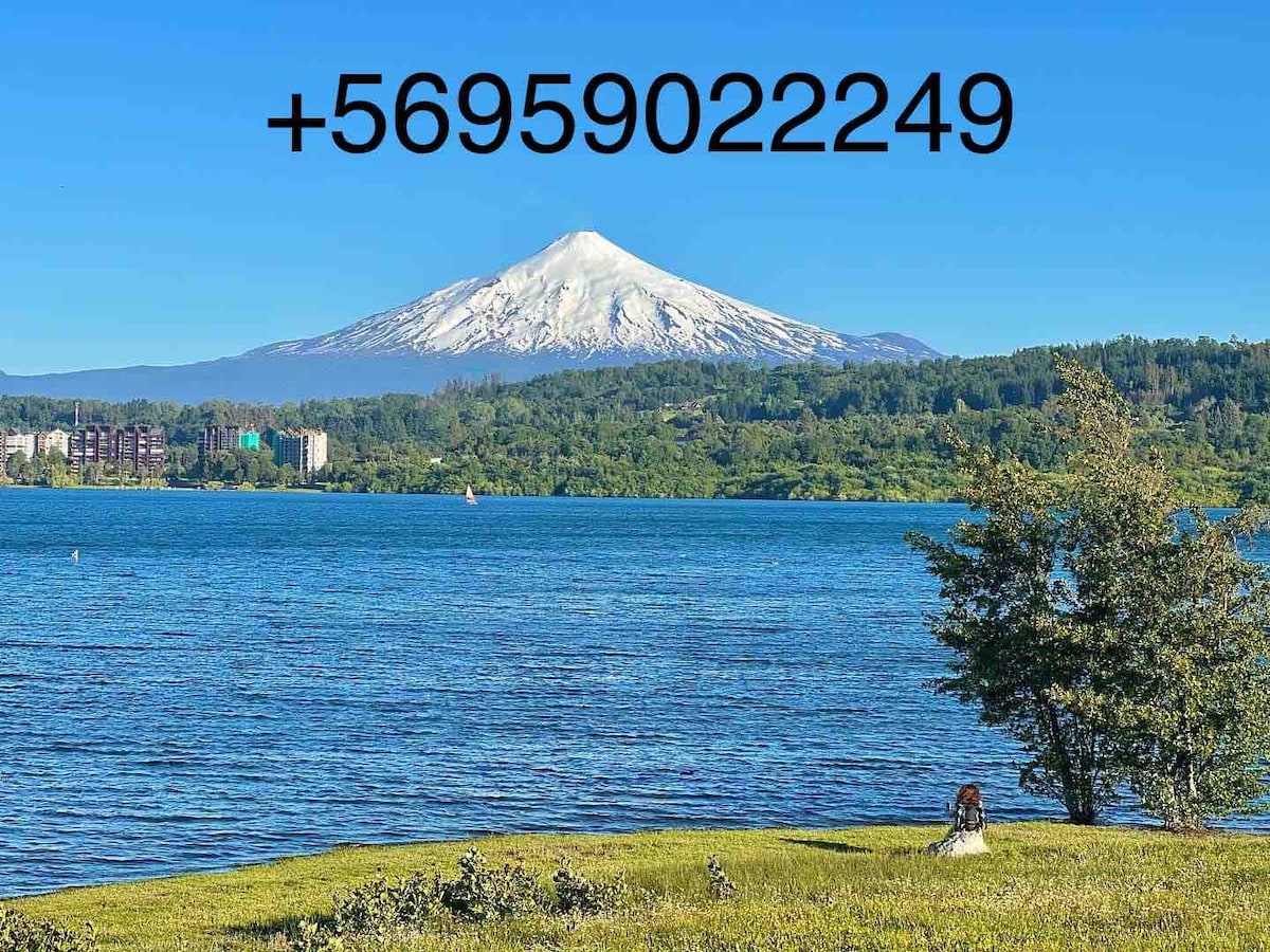 A serene lakeside view showcases a snow-capped mountain in the distance, surrounded by lush greenery. A calm body of water reflects the mountain, while a person sits peacefully on the grassy shore, enjoying the natural landscape.
