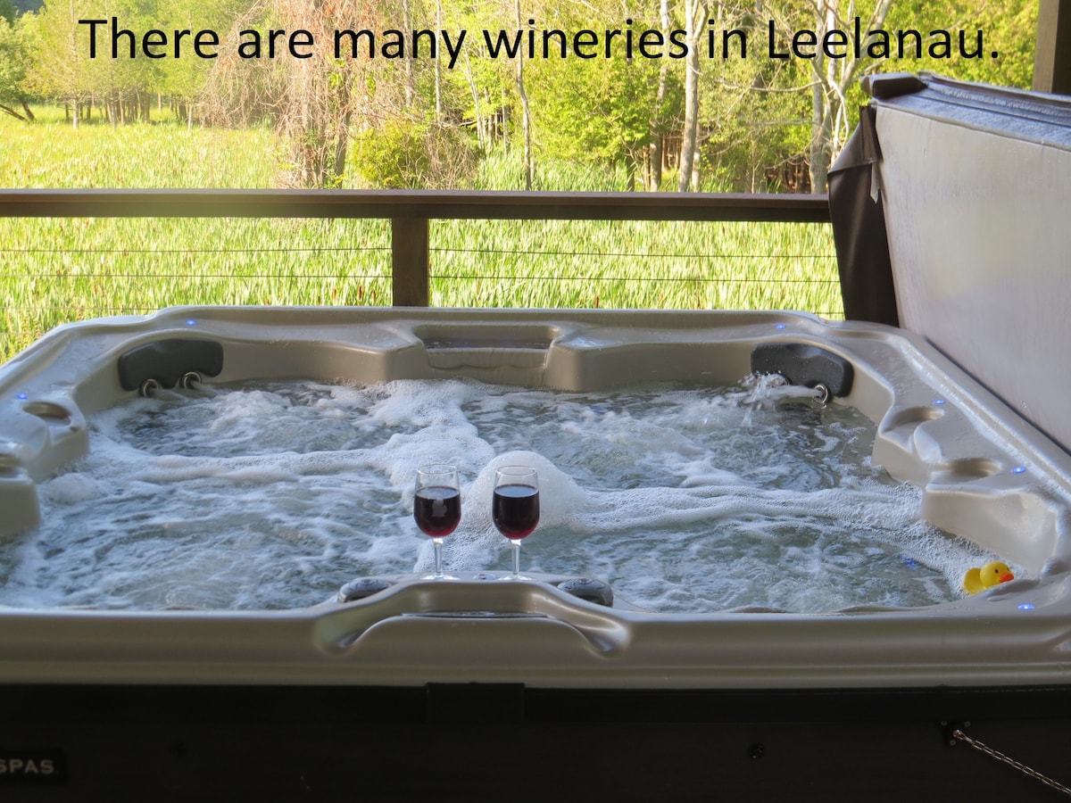 A spacious hot tub is displayed on a covered deck, surrounded by a wooden railing and greenery. Two glasses of red wine are set on the edge of the tub, with bubbles visible in the water, suggesting a relaxing atmosphere.