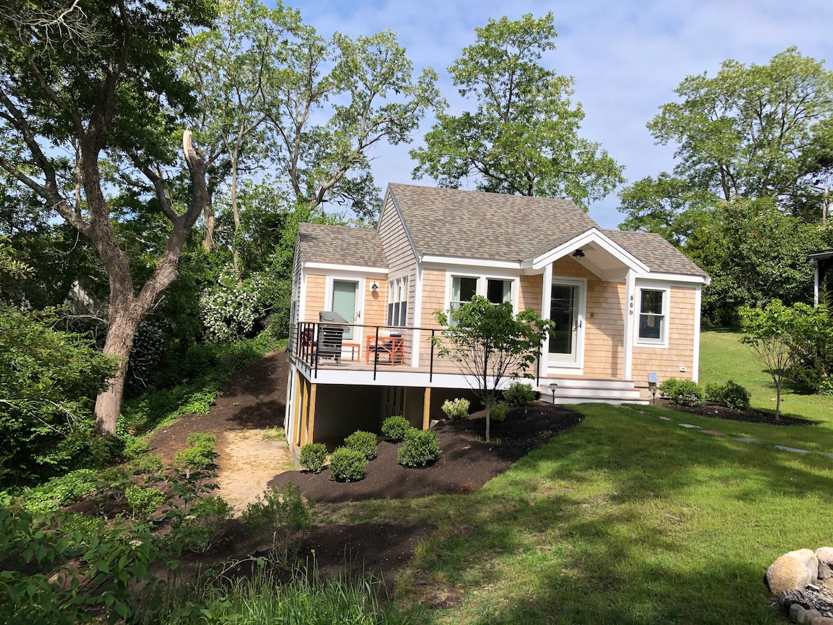A charming cottage is nestled on a hillside, surrounded by lush greenery. The exterior features a light wood facade and a welcoming front porch with seating. The landscaped yard is manicured, offering a natural setting that blends harmoniously with the surrounding environment.