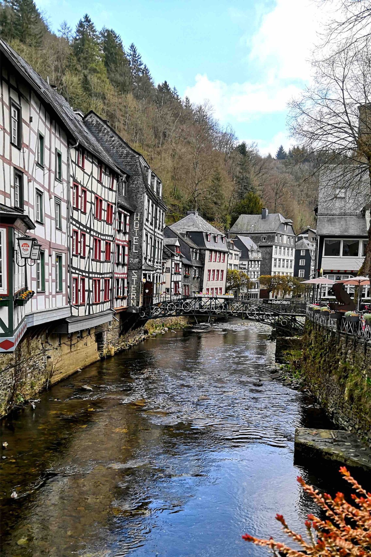 Historic buildings line both sides of a gently flowing river, reflecting the architectural charm of the area. Natural greenery is visible in the background, complementing the serene atmosphere. The scene captures a blend of nature and history, highlighting the quaint appeal of the surroundings.
