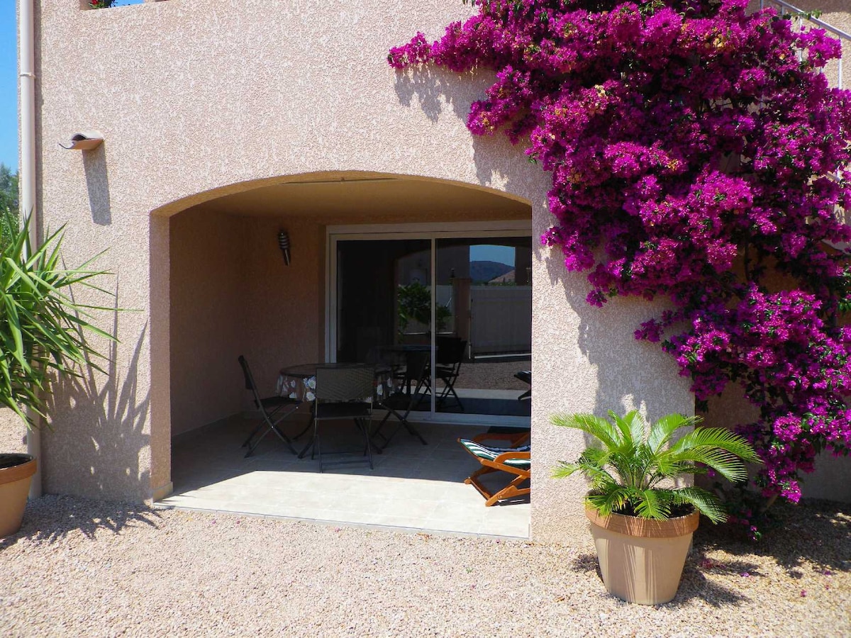 An outdoor seating area is framed by vibrant bougainvillea, creating a natural backdrop. A table with chairs sits on a tiled surface, inviting relaxation. Potted plants add greenery, while large glass doors provide a view of the interior, blending indoor and outdoor spaces.
