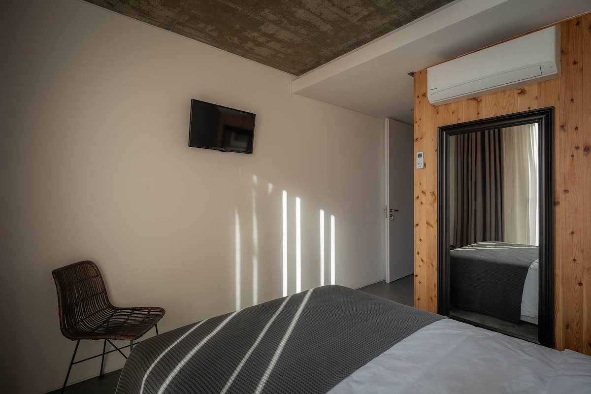 A minimalist bedroom features a spacious bed covered with a textured gray blanket. A wire chair is positioned nearby, and a wall-mounted television is visible. Natural light streams through a window, casting patterns on the wall, while a mirror reflects the room's neutral color palette.