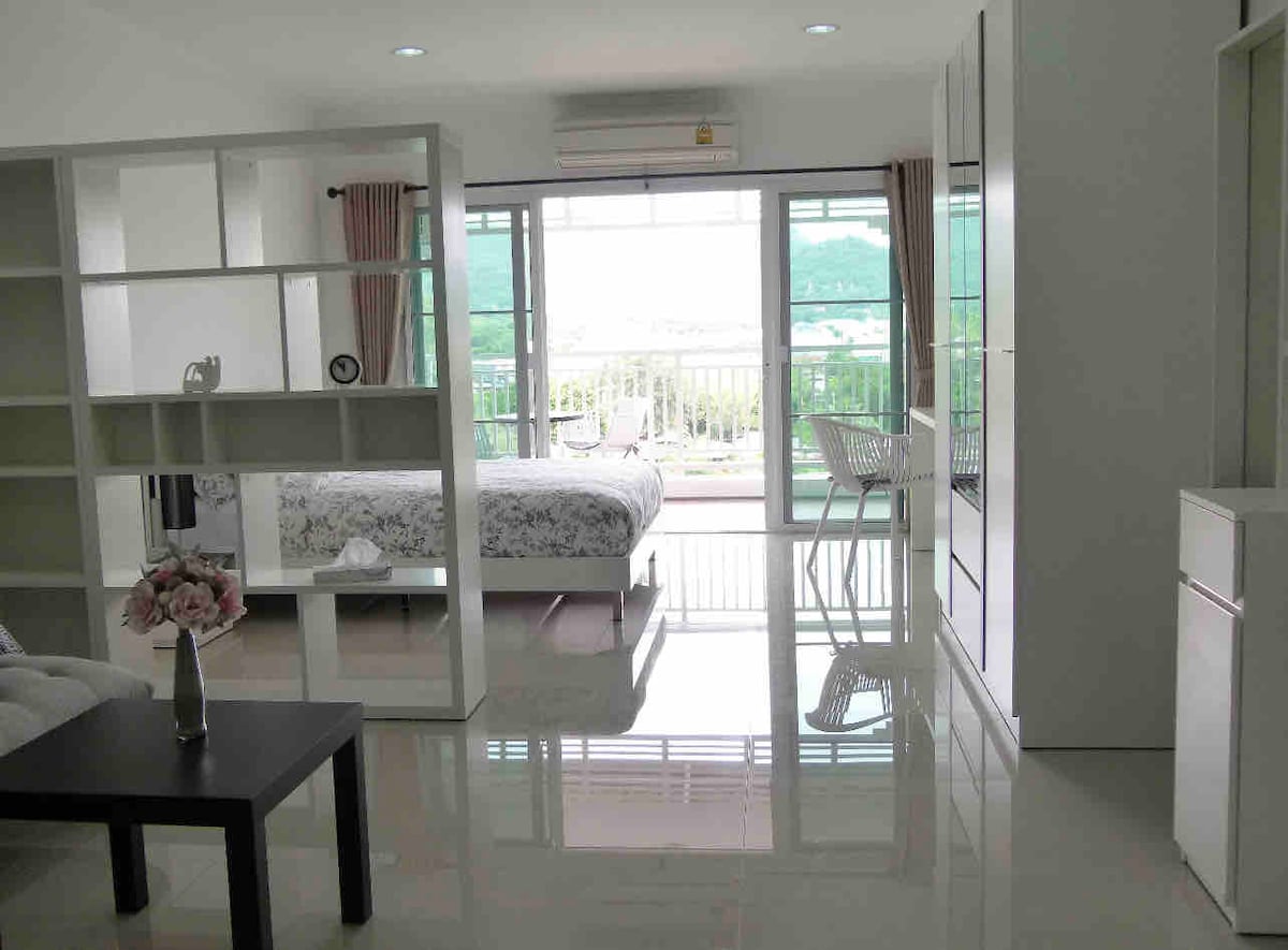A spacious studio apartment features a bright living area with large windows allowing natural light to fill the space. The modern furnishings include a cozy bed, a dining table with chairs, and a stylish coffee table. A private balcony with scenic views of the mountains is visible.