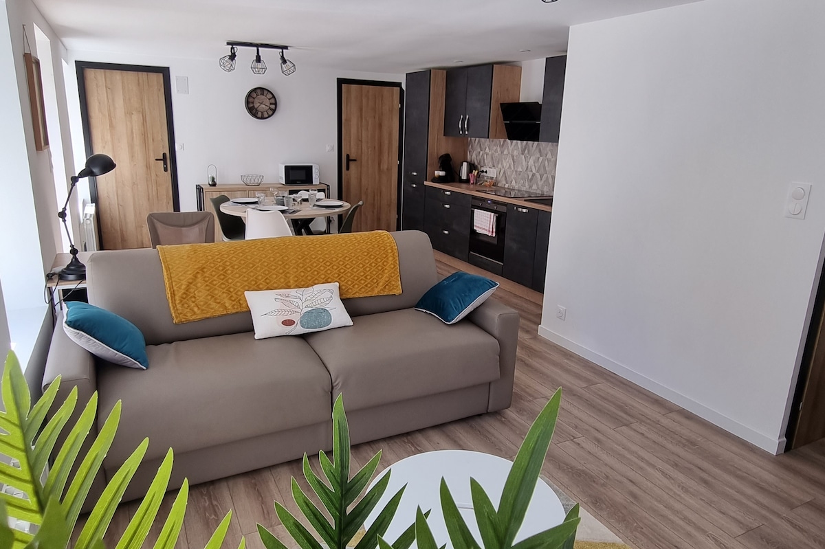 A living area is presented, featuring a neutral-toned sofa adorned with colorful cushions. A dining table is positioned nearby, accompanied by modern dining chairs. The kitchenette is visible, showcasing dark cabinetry and a stylish backsplash. Natural light fills the space through large windows.