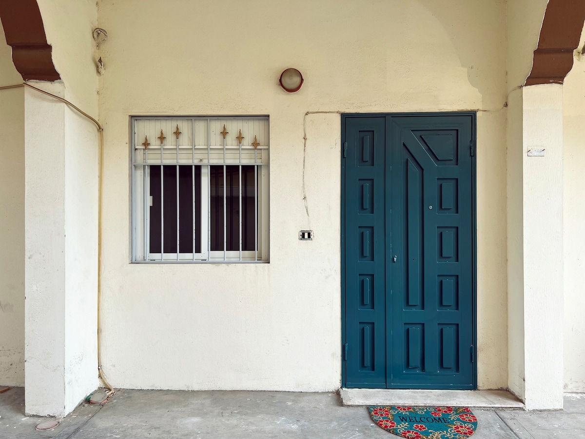 The exterior features a private entrance with a green door and a decorative welcome mat. A window secured with metal bars is present beside the entrance, providing natural light. The area is well-maintained, with a simple concrete floor visible.