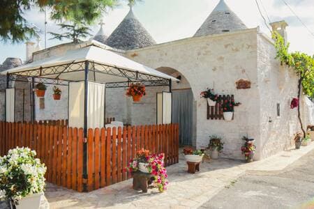 An outdoor gazebo is surrounded by a wooden fence and flowering plants, providing a cozy area for relaxation. The charming trulli structure features stone walls and traditional conical roofs, enhanced by hanging flower pots that add a touch of vibrancy to the setting.