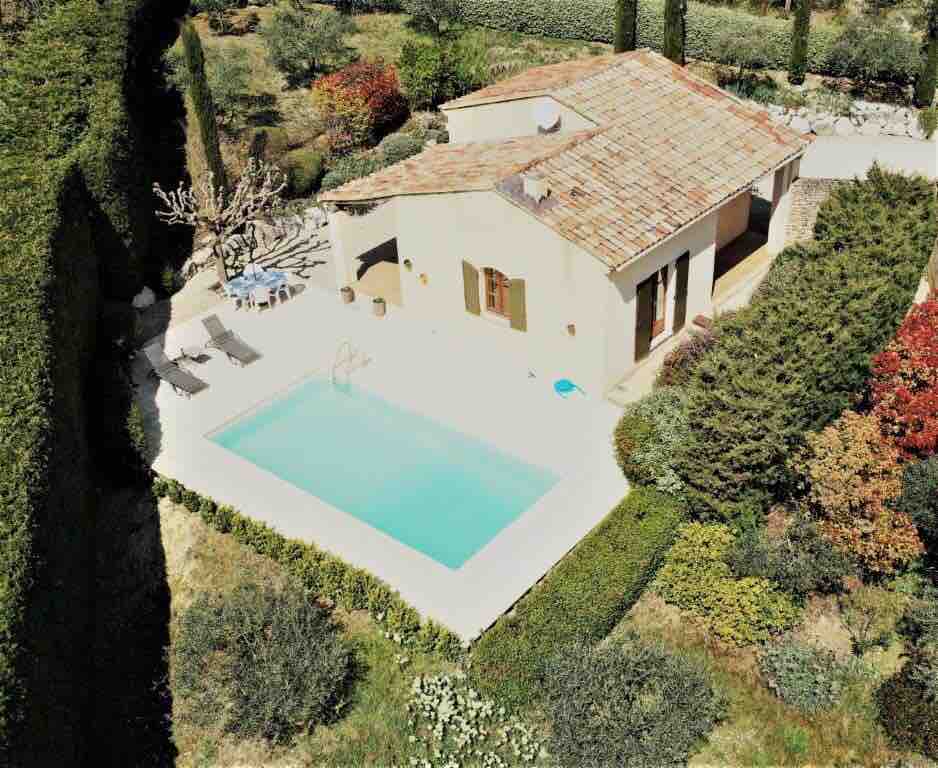 An aerial view of the property reveals a private pool surrounded by a landscaped garden. The villa features a terracotta roof and is framed by neatly trimmed hedges. Outdoor lounge chairs are positioned beside the pool, inviting relaxation and enjoyment of the scenic surroundings.