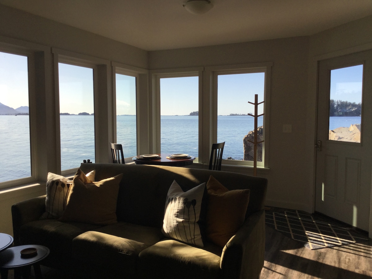 A cozy living space features a comfortable sofa adorned with decorative pillows. Large windows provide a panoramic view of the calm ocean and distant islands, filling the room with natural light. A small table is set for two, enhancing the inviting atmosphere.