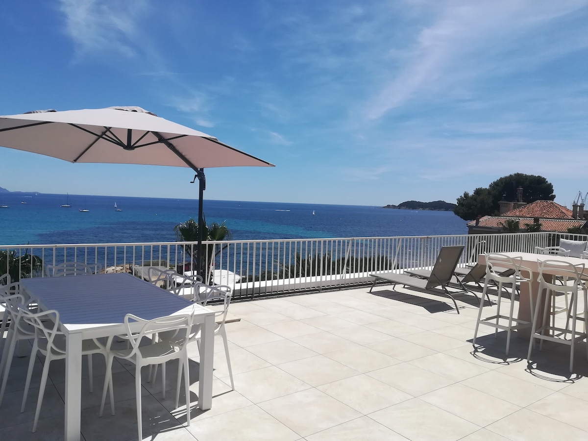 An expansive private terrace is featured, designed for outdoor relaxation and dining. It includes a large table surrounded by chairs and several lounge chairs. A large umbrella provides shade, and stunning views of the sea and sailboats are visible in the background.