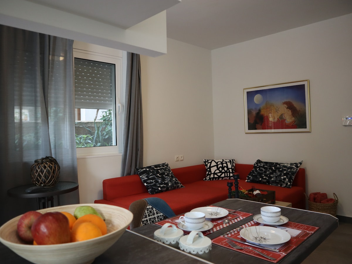 A modern living area features a bright red sofa adorned with patterned cushions. A dining table is set with white dishware and a fruit bowl containing apples and oranges. Natural light enters through a window, which is partially covered by sheer curtains.