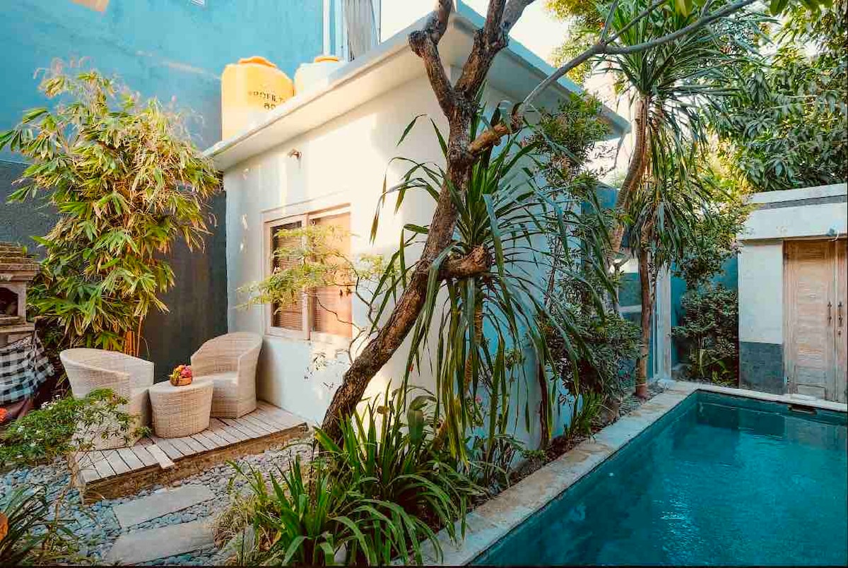 A serene outdoor space features two wicker chairs arranged beside a small wooden deck, surrounded by lush greenery. A tranquil pool reflects the surrounding plants while a small building is visible in the background, contributing to a peaceful natural setting.