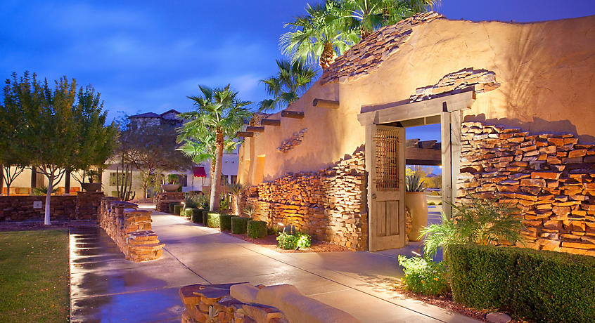 A stone pathway leads through lush landscaping, with desert plants and palm trees flanking both sides. The rustic stone wall features a wooden entry gate, softly illuminated in the evening light, creating a welcoming ambiance.