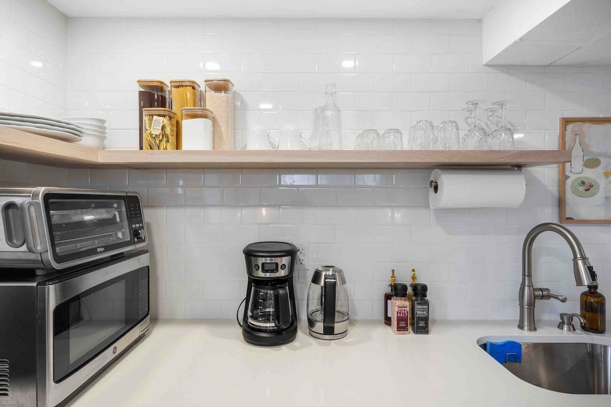 The kitchenette is equipped with modern appliances including a microwave, coffee maker, and kettle. Storage containers, glassware, and dinnerware are neatly arranged on open shelves. A paper towel roll is conveniently placed above the sink, enhancing functionality in the compact space.