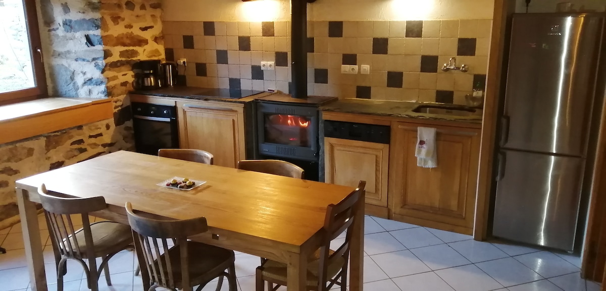 A spacious kitchen area showcases a wooden dining table surrounded by six chairs. A fireplace is positioned between the cabinetry and appliances, which include an oven, microwave, and refrigerator. Natural light enters through a large window, highlighting the stone walls and tiled floor.