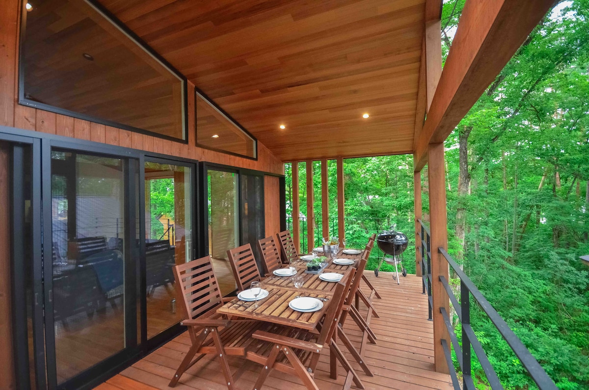An outdoor deck features a long wooden table with seating for ten, surrounded by lush greenery. Large glass doors provide a view into the interior. A BBQ grill is positioned nearby, enhancing the space for outdoor dining and activities.