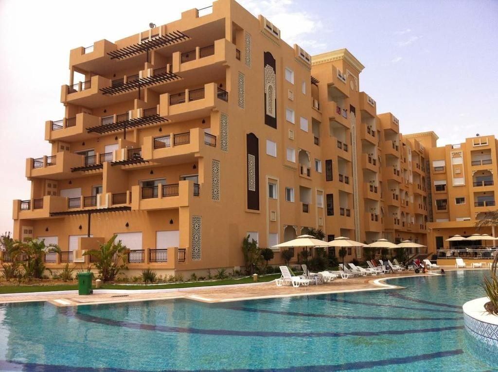 A modern apartment building features multiple balconies with decorative railings. Surrounded by a serene pool area, the structure displays a palette of warm earth tones. Sun loungers are arranged near the water, and lush greenery enhances the inviting setting.