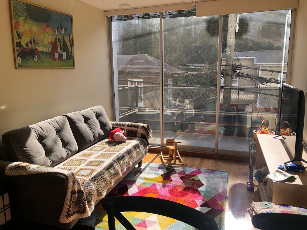 A bright living space is filled with natural light, showcasing a comfortable couch adorned with a colorful blanket. A vibrant rug adds warmth to the wooden floor, while a large glass door leads to a balcony. A TV and playful items are situated along the wall.